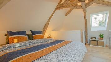 Ein Schlafzimmer mit schrägen Wänden, einem großen Bett mit gemusterten Betttextilien, einem Nachttisch mit Lampe und einem Fenster mit Blick auf Bäume.