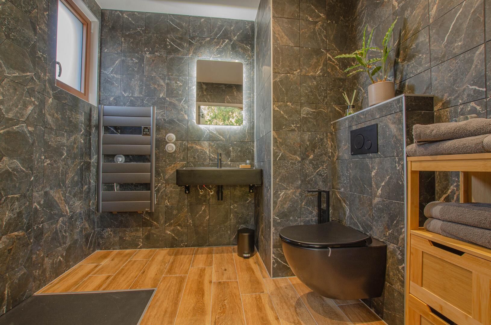 A modern bathroom with a marble wall, a wooden floor, a black toilet, a sink, and a towel rack.