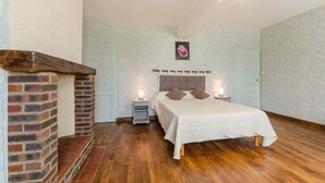 Un dormitorio con chimenea de ladrillo, una cama con cabecera, dos mesitas de noche con lámparas, y una puerta al fondo.
