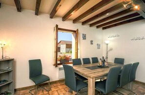 A dining area with a wooden table and chairs, a bookshelf, and a window with a view of a building and flowers outside.