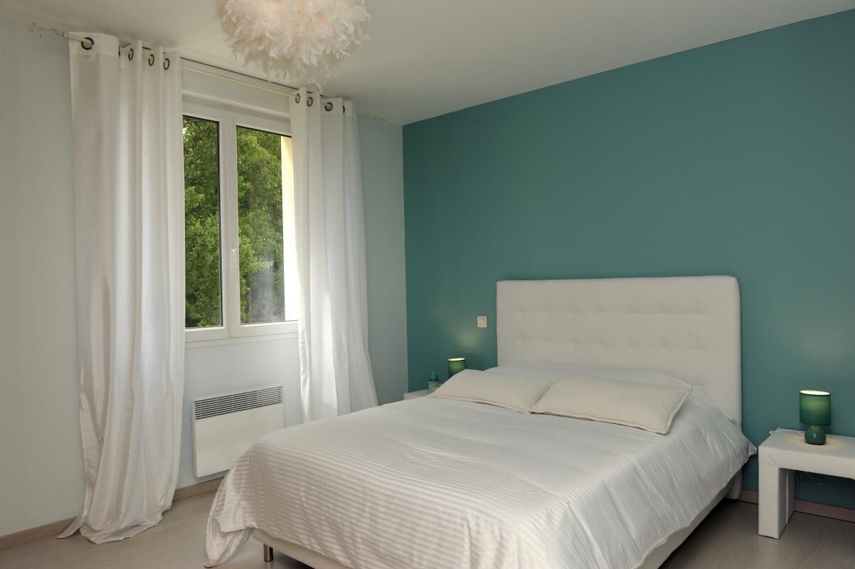 A bedroom with a bed, white curtains, a window, and a nightstand with a lamp.