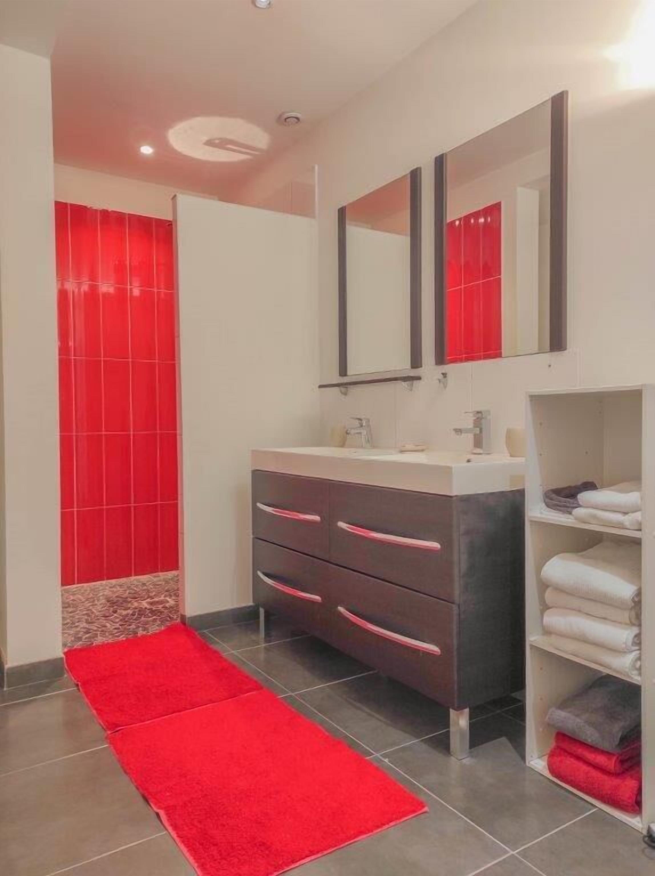 Un baño moderno con zona de ducha revestida en mosaicos rojos, un lavabo blanco con espejo y una alfombra roja.