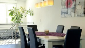 A dining area with a table, chairs, a glass of beverage, a hanging light fixture, and a wall-mounted artwork.