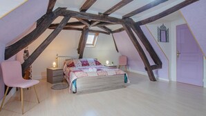 Et soveværelse med skråt loft, synlige træbjælker, en seng med quilt, en stol, sengeborde og en dør.