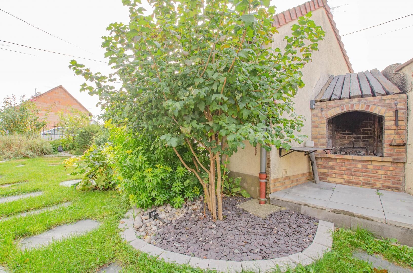 Un jardin avec un espace barbecue en briques, un petit arbre et un chemin en pierre.