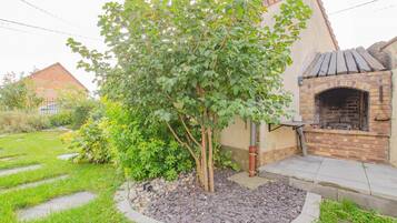 Un jardin avec un espace barbecue en briques, un petit arbre et un chemin en pierre.