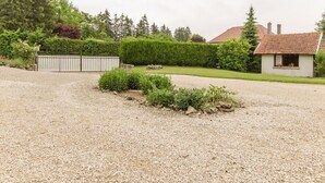 Une allée de gravier mène à une maison dont le toit est recouvert de tuiles rouges, et qui est entourée d’un jardin bien entretenu et d’une clôture en métal.