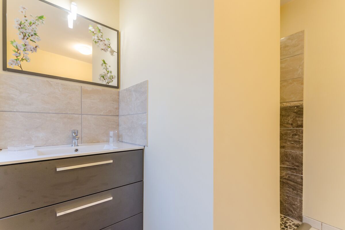 Une salle de bain avec un lavabo blanc, des tiroirs foncés, un miroir orné de motifs floraux et un mur de douche en pierre.