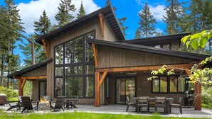 Una casa moderna de dos pisos con amplios ventanales, un patio con mobiliario exterior y un bosque de fondo.