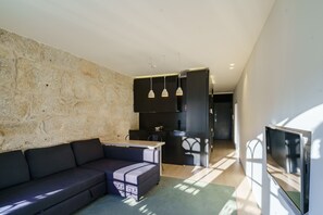 A modern living space with a stone wall, a dark sofa, a kitchen area with black cabinets, and a flat-screen TV. - N Apartments - Chã 101 (Porto)