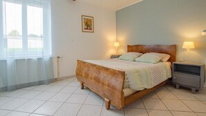 Un dormitorio con una cama de madera, dos mesitas de noche con lámparas, un radiador y una ventana con cortinas translúcidas.