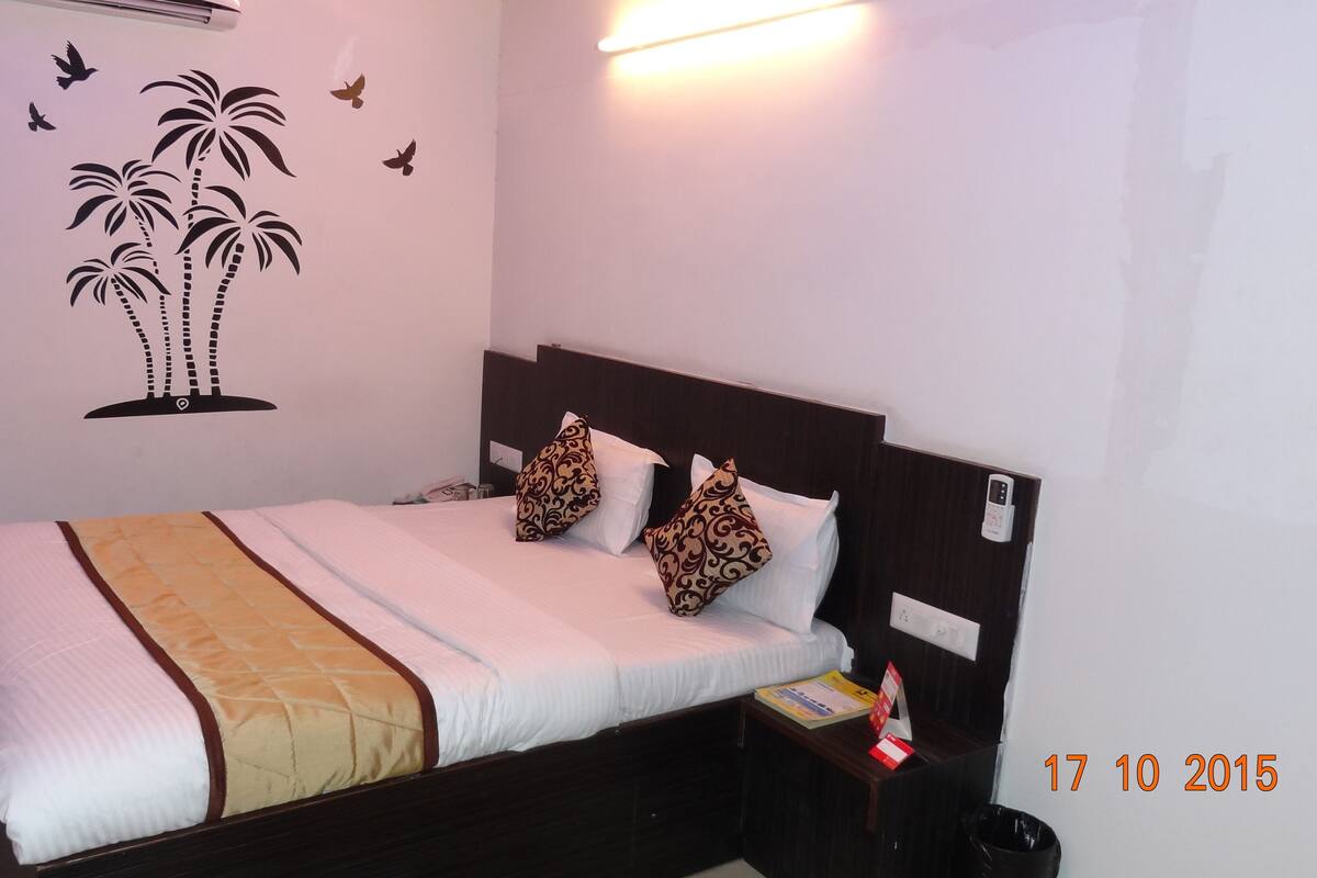 A hotel room with a bed, two decorative pillows, a nightstand, a wall-mounted air conditioner, and a wall decal of palm trees and birds.