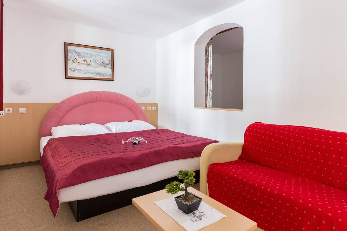 A bedroom with a bed, a red sofa, a small table with a plant, a framed picture on the wall, and a mirror above the sofa.