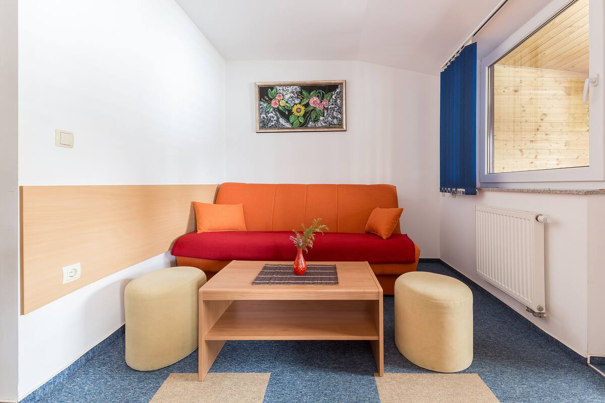 A small, cozy room with a red sofa, a wooden coffee table with a vase, and a window with wooden blinds.