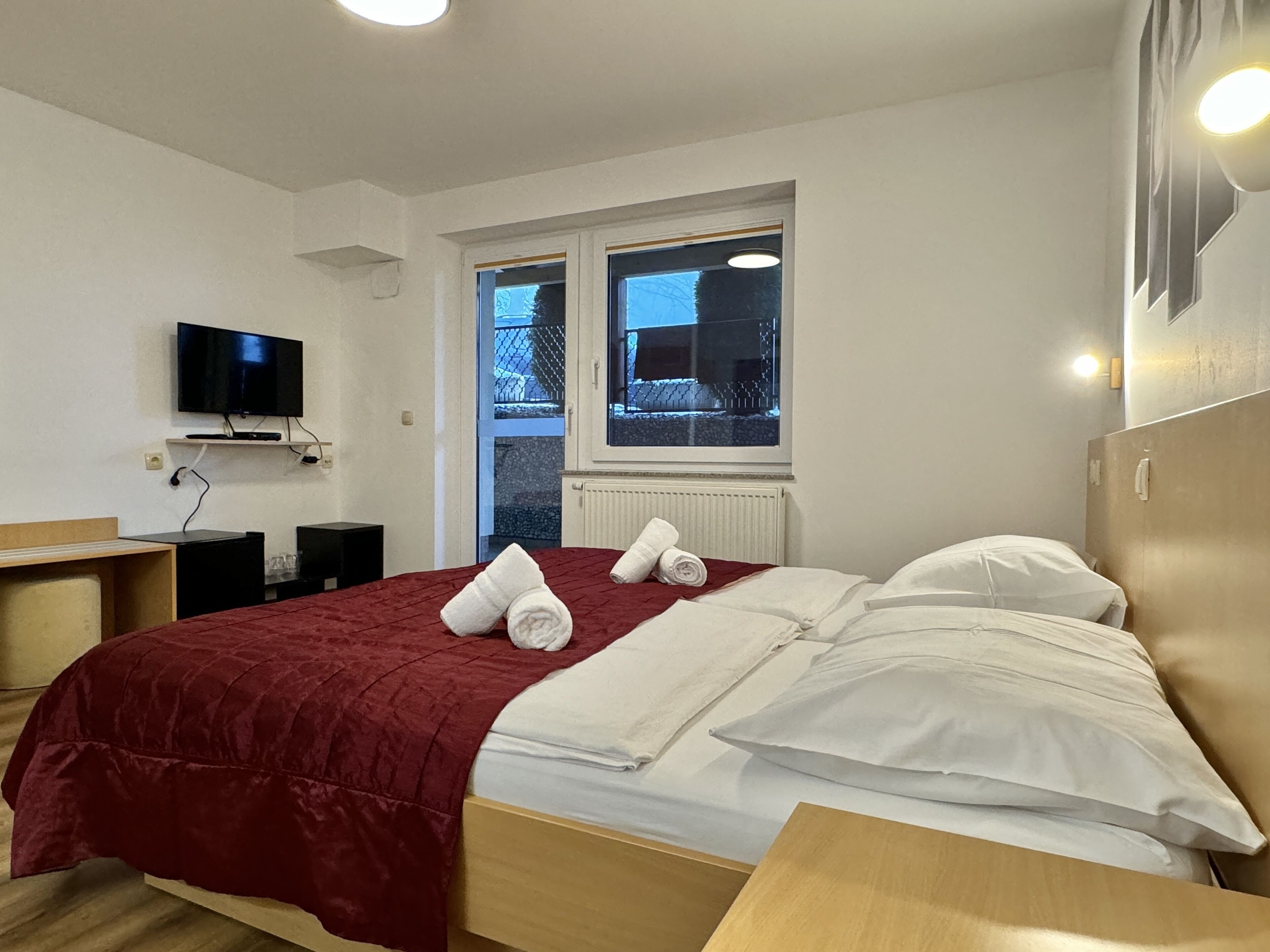 A hotel room with a bed, a TV mounted on the wall, a window with a view of the outside, and a radiator under the window.