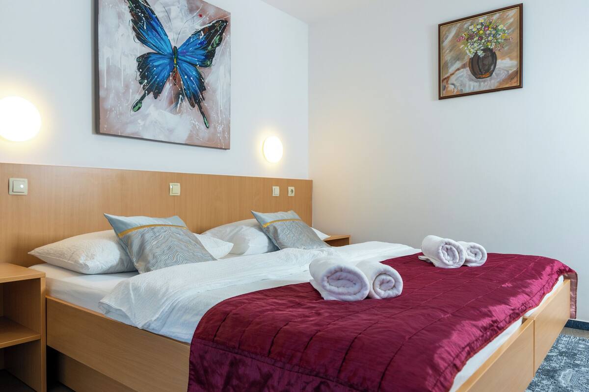 A neatly made bed with a red blanket, pillows, and rolled towels. There is a wooden headboard, a nightstand, and a wall-mounted painting of a blue butterfly.