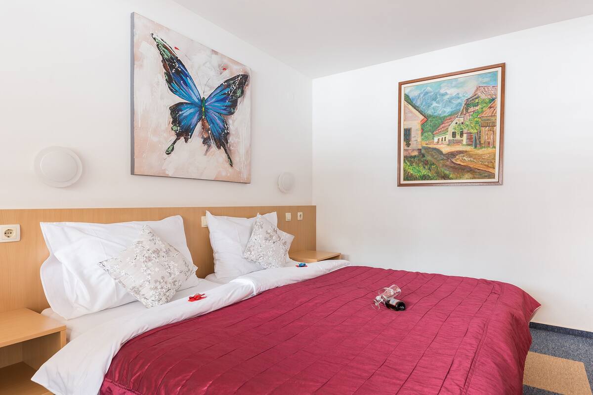 A hotel room with a bed, white bedding, a red blanket, a painting of a butterfly, and a landscape painting.
