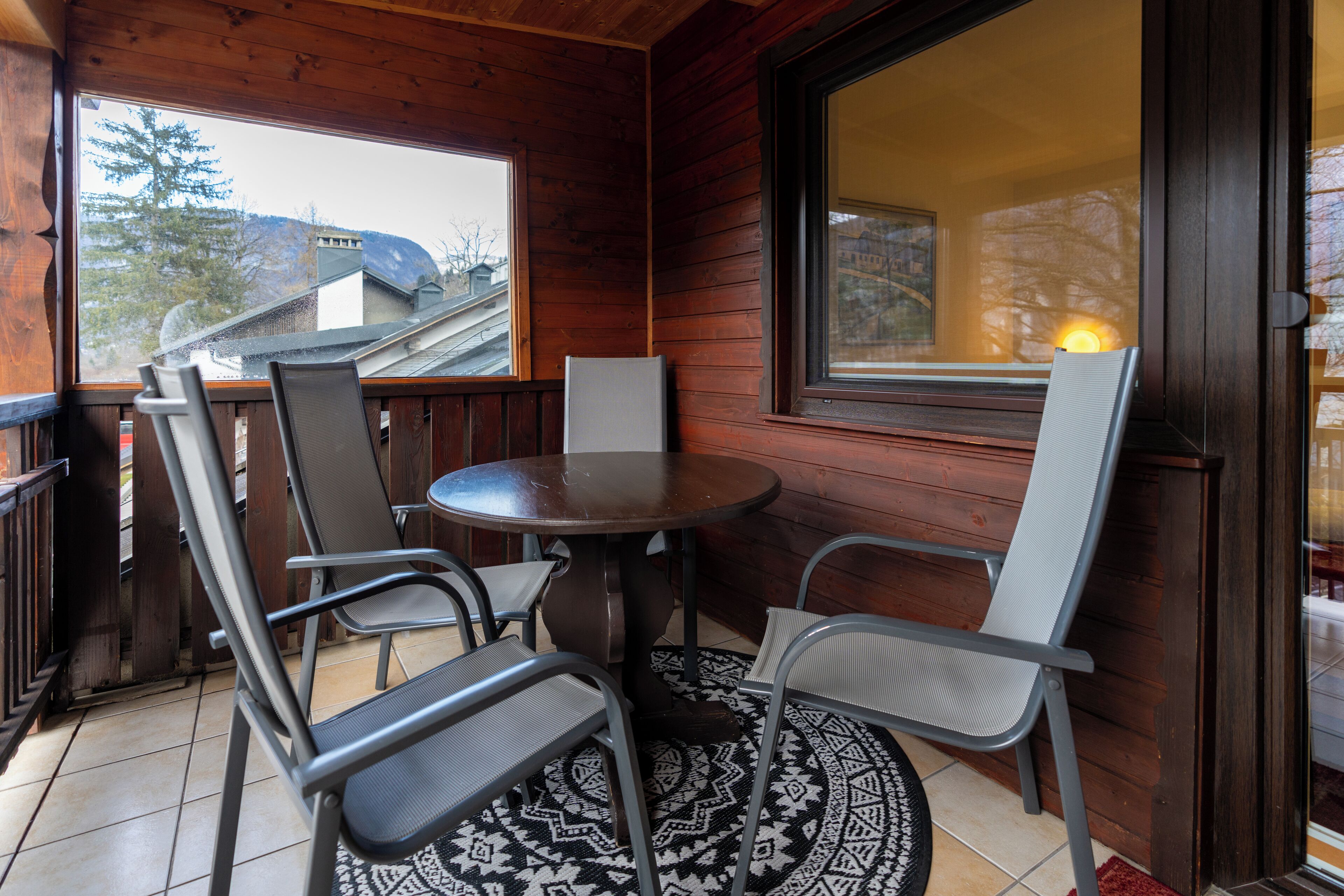 Um deck de madeira com uma mesa redonda e quatro cadeiras, uma janela grande e vista para edifícios e árvores.