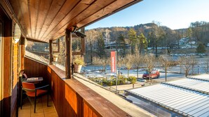 A wooden balcony with a view of a parking lot and a building across a river. - Hotel Center (Bohinj)