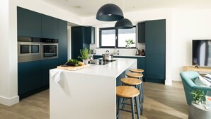 A modern kitchen with dark teal cabinets, stainless steel appliances, a central island with stools, and a large window providing natural light. - Luxury Newquay home with epic views - dog friendly (Newquay)