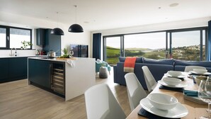 A modern kitchen with a central island, dining area with white chairs, and a large window offering a view of the countryside. - Luxury Newquay home with epic views - dog friendly (Newquay)