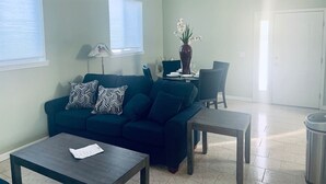 Una sala de estar con un sofá azul oscuro, una mesa de centro, una mesa de comedor con sillas, un ventilador de techo y un florero con flores.