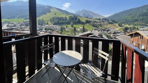 A balcony with a table and chair, overlooking a town with mountains in the background. - RESIDENCE 2-234 - Village center apartment, close to the slopes - Wi-Fi - Garage (La Clusaz)
