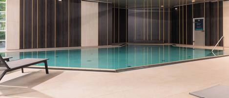 An indoor swimming pool with clear water, surrounded by walls with vertical lines, and equipped with lounge chairs and a table.