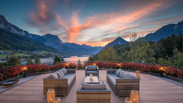 A deck with wicker furniture and candles, overlooking a mountainous landscape and a lake.