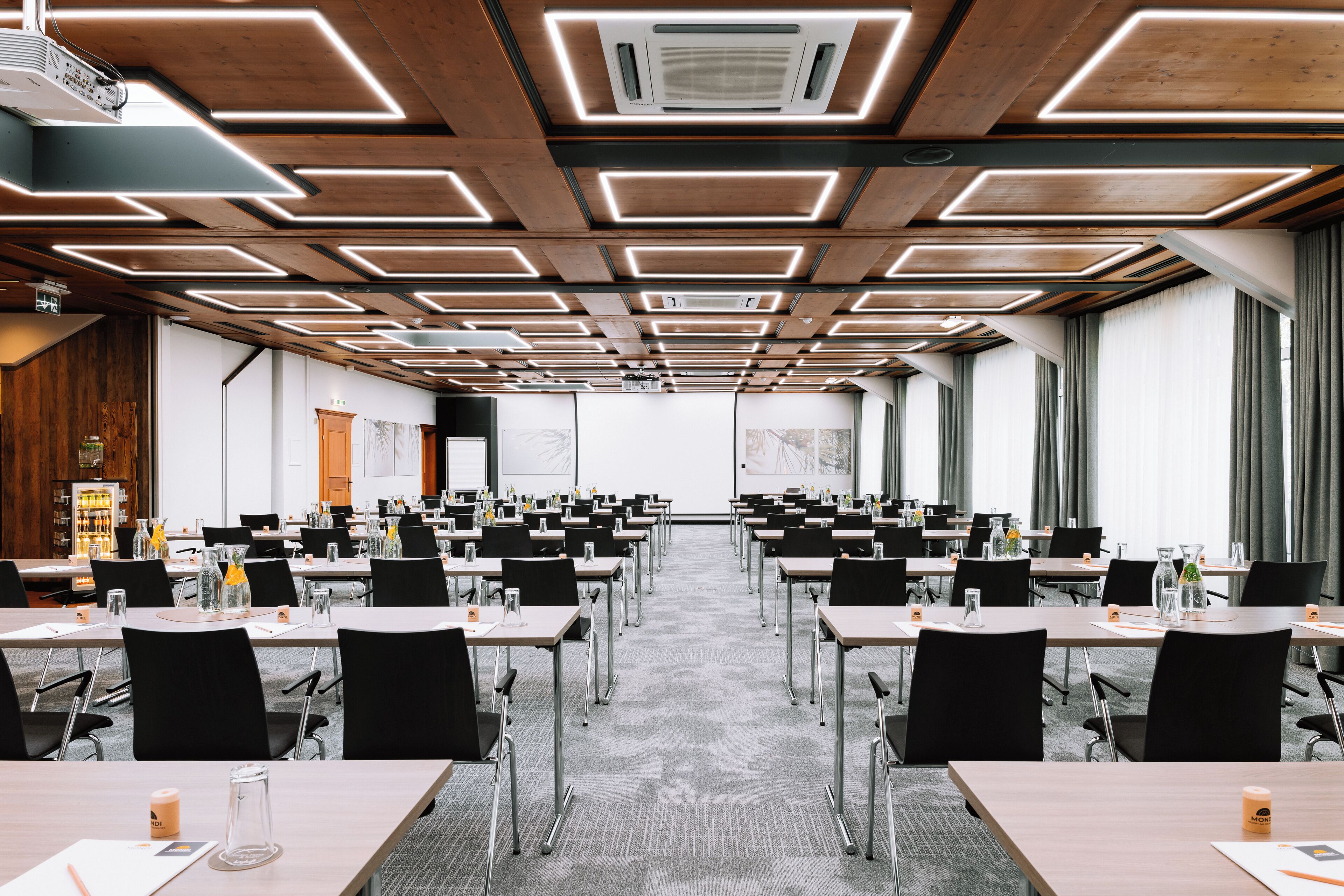 Una sala de conferencias con varias mesas y sillas, techo de madera con iluminación geométrica y ventanas amplias con cortinas.