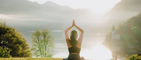 A person meditating on a grassy area with a lake and mountains in the background.