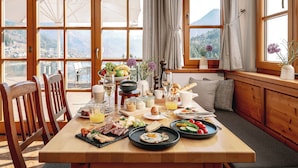 A breakfast table with various foods and drinks, wooden chairs, and large windows with curtains.