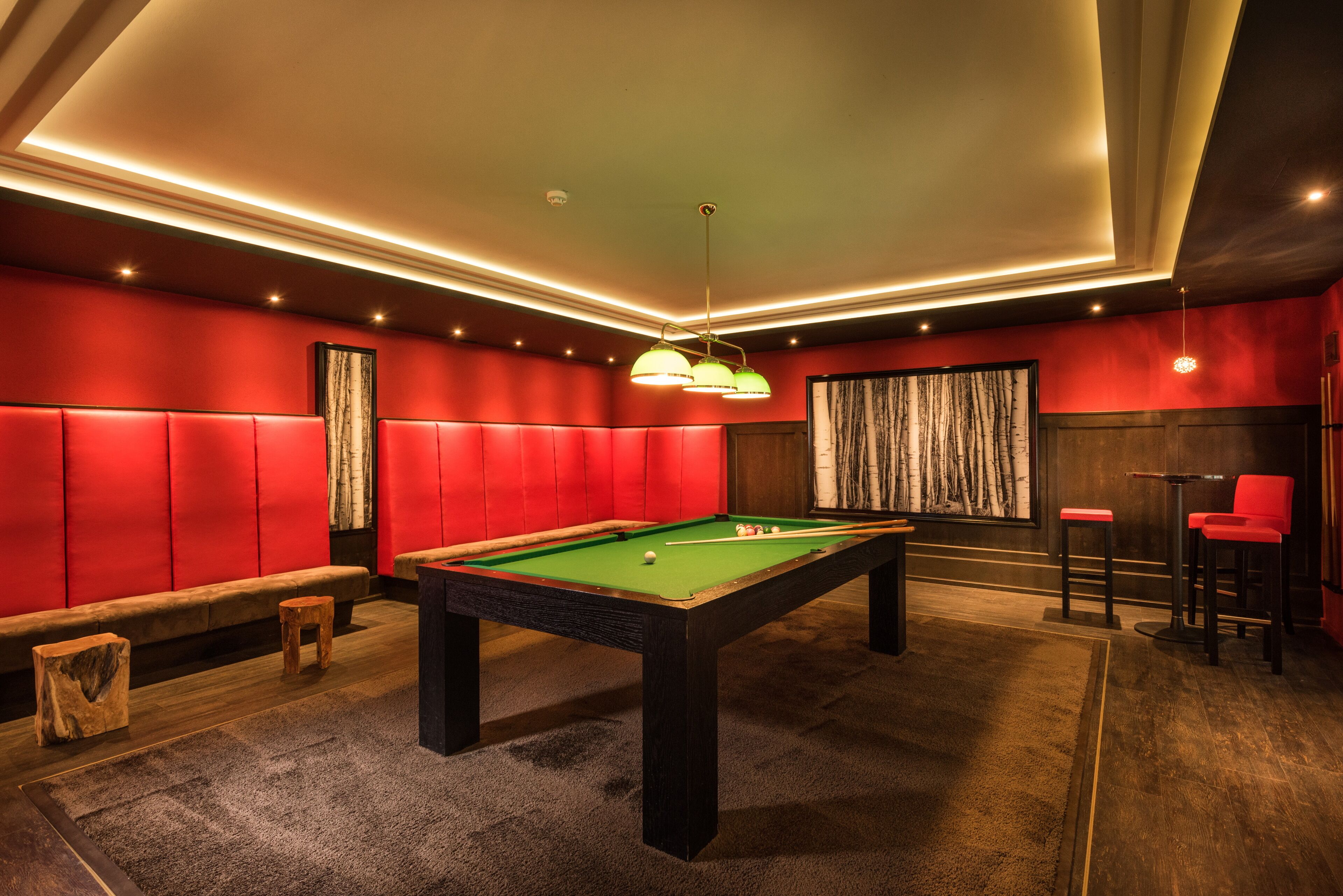 A pool table in a room with red padded walls, a bench, and a bar area with red chairs.
