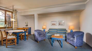 A room with a dining table and chairs, a sofa, two armchairs, a small table with a lamp, and a window with curtains.
