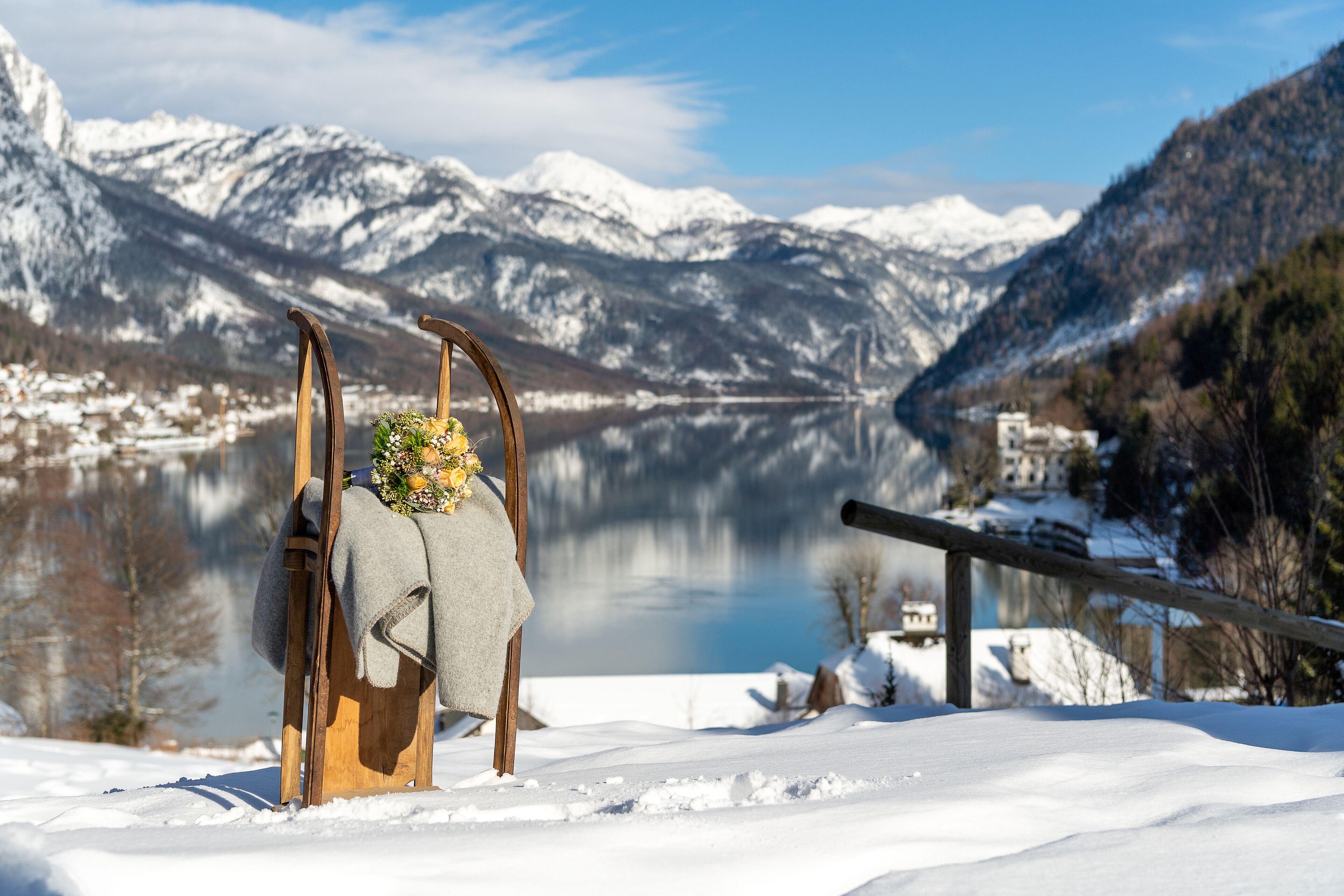 A wooden chair with a blanket and a floral arrangement in a snowy landscape with a lake and mountains in the background.