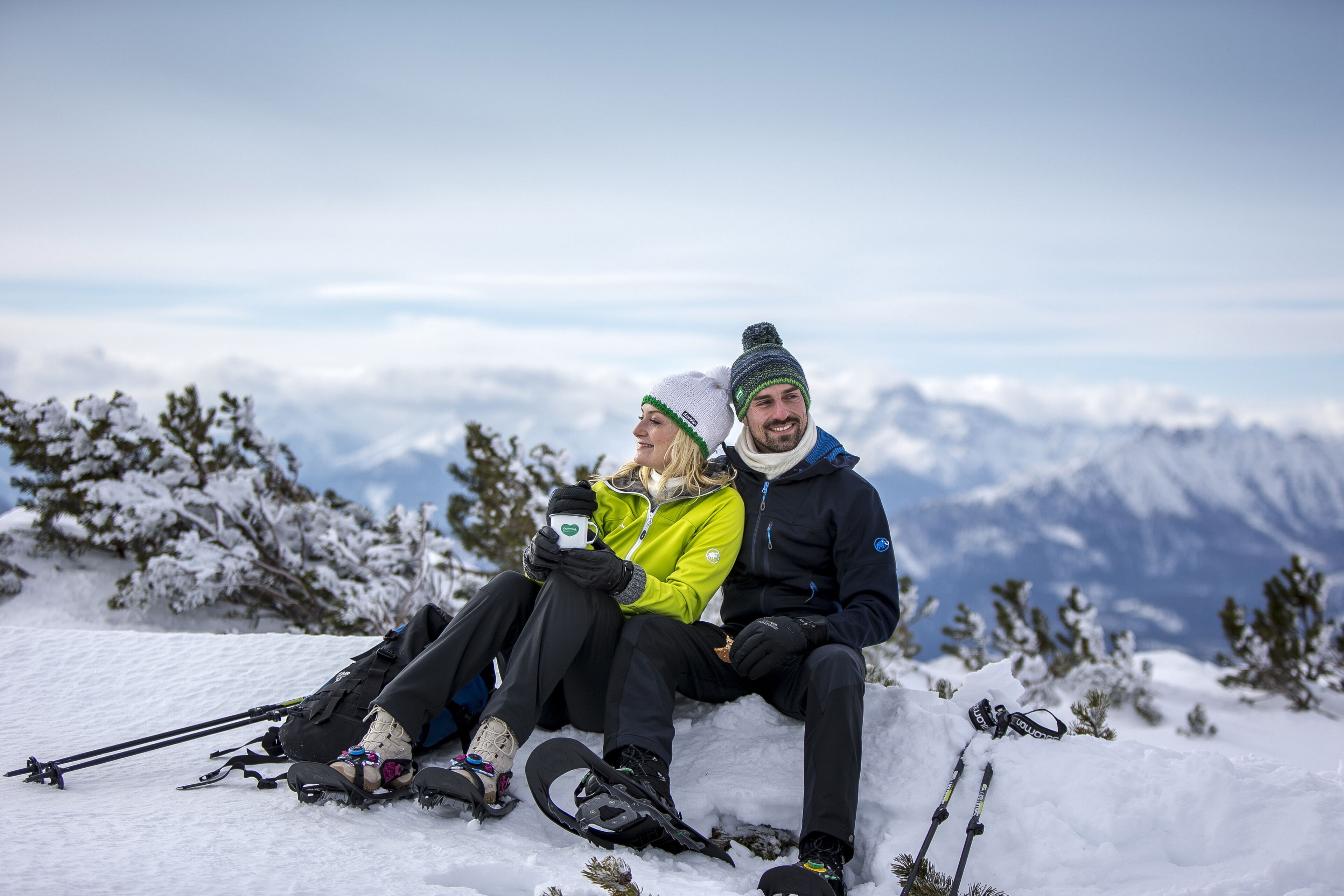 A couple sitting on a snow-covered slope with a mountainous backdrop.