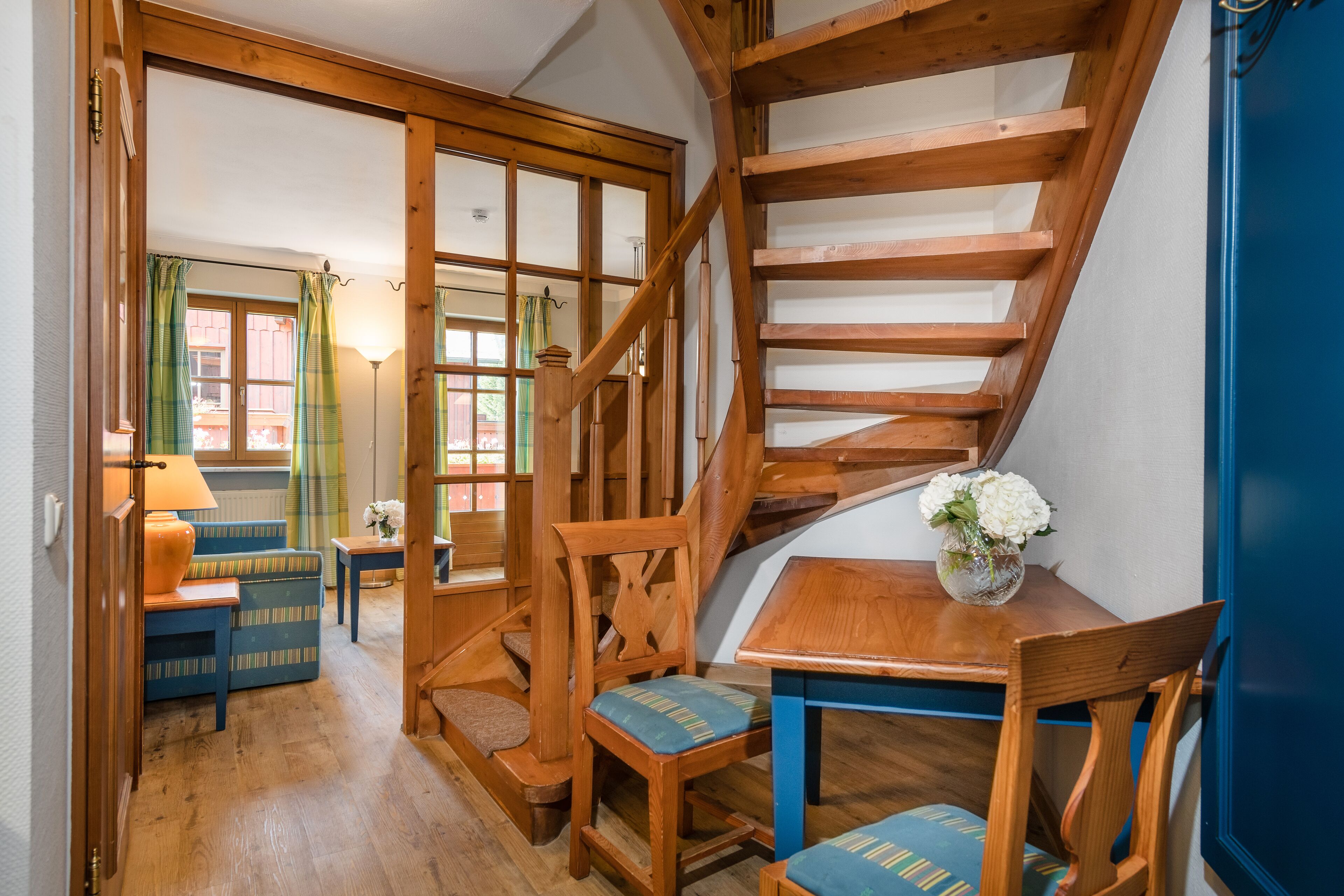 Una escalera de madera que lleva a un piso superior, una mesa de comedor de madera con sillas, un sofá y una lámpara en una habitación con piso de madera y vista por una puerta de vidrio.