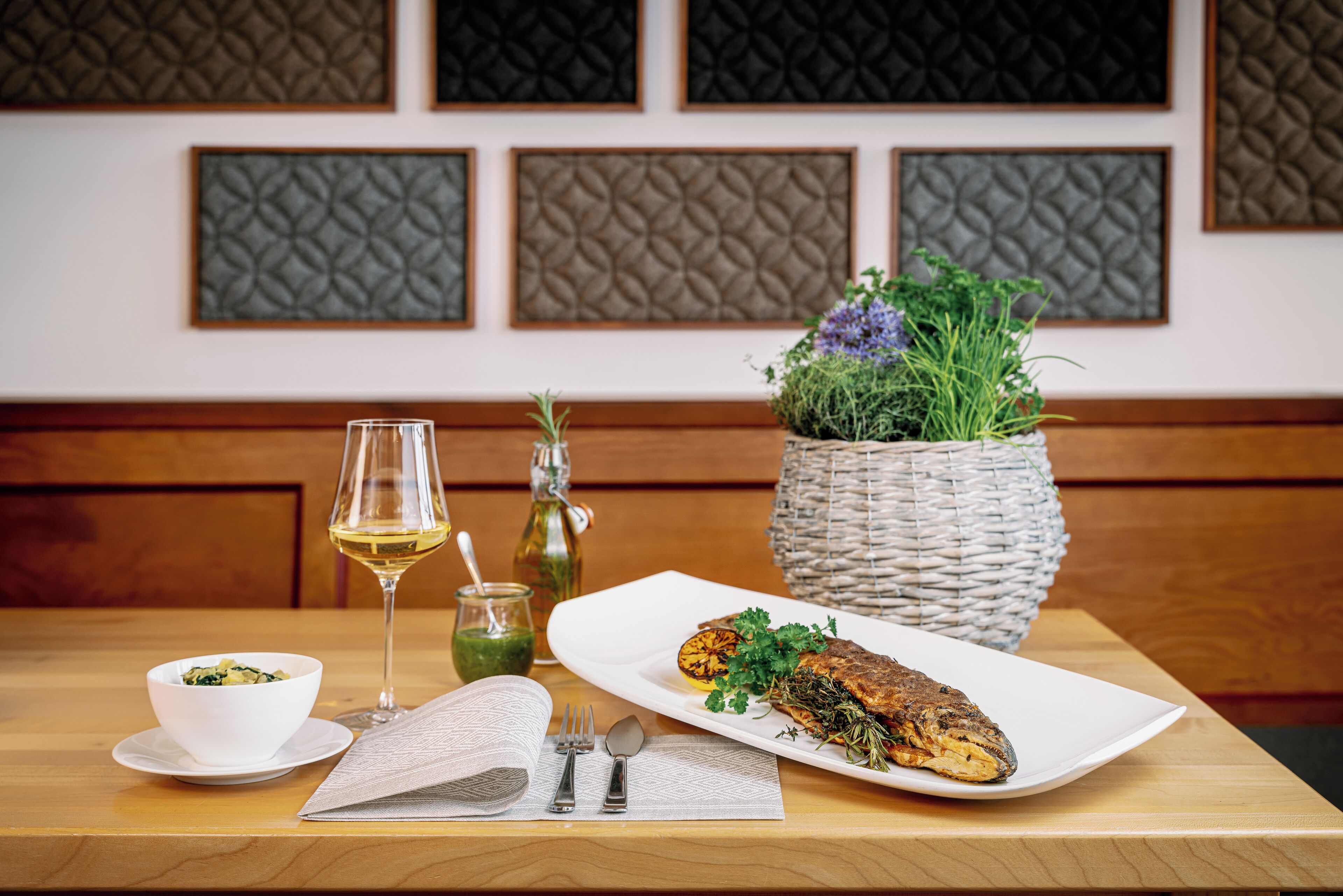 Une table à manger avec une assiette blanche contenant du poisson, un saladier, un verre à vin et un panier tressé rempli de verdure.
