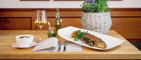 Una mesa de comedor con un plato de pescado blanco, un bol de ensalada, una copa de vino y una cesta tejida con follaje.
