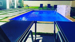 A swimming pool with lounge chairs and a covered area with doors and air conditioning units.