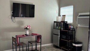 Een compacte woonruimte met een flatscreen televisie aan de muur, een kleine tafel met stoelen, een koelkast, een afvalbak en een raam met zonwering.