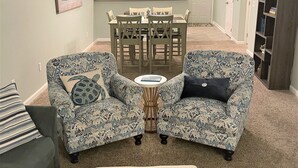 Ein Wohnzimmer mit zwei gemusterten Sesseln, einem Couchtisch mit Tablett, einem Essbereich mit Tisch und Stühlen sowie einer Küche im Hintergrund.