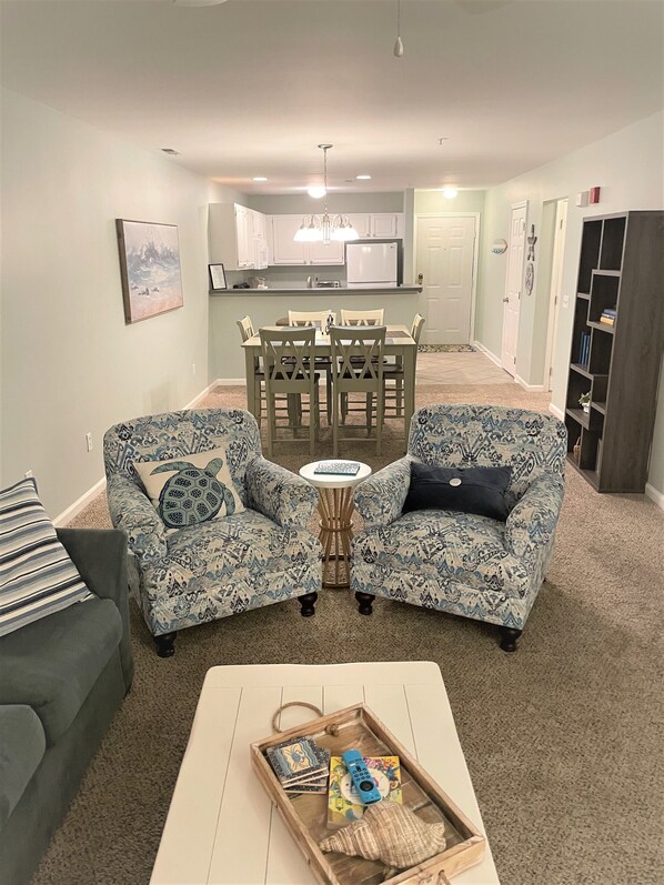 A living room with two patterned armchairs, a coffee table with a tray, a dining area with a table and chairs, and a kitchen in the background.