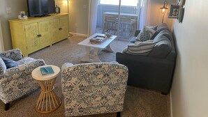 Ein Wohnzimmer mit einer Couch, Sesseln, einem Couchtisch, einem Fernseher auf einem gelben Schrank und Blick durch eine Glastür.