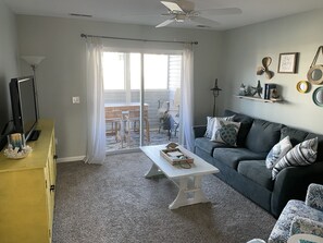 A living room with a dark grey sofa, a white coffee table, and a yellow cabinet. There is a television on the left and a ceiling fan above.