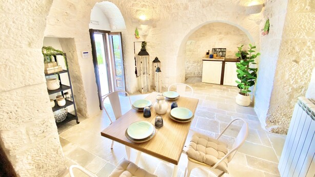 Interior rústico de piedra con una mesa de comedor preparada para cuatro personas, rodeada de muros de piedra y una cocina visible al fondo.