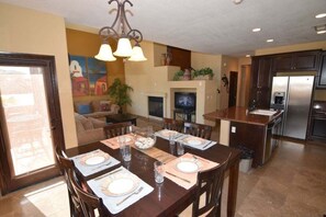 Uma sala de jantar com uma mesa posta para quatro pessoas, uma cozinha equipada com frigorífico e uma sala de estar com lareira.