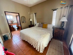 Un dormitorio con una cama grande, una mesita de noche con lámpara, una ventana con cortinas y un baño a simple vista al fondo.