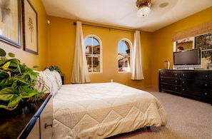 Une chambre à coucher comprenant un lit, une commode, un téléviseur et deux fenêtres avec des rideaux.