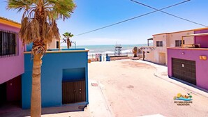 A narrow street with buildings on either side, a palm tree, and a beach in the background.